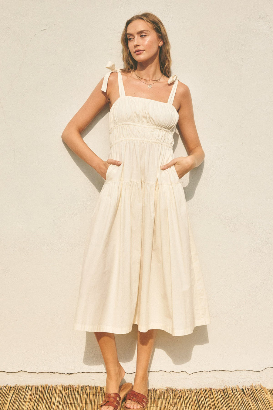Featuring an adjustable tie strap flare midi dress with a square neck and sinching along the middle and pockets on each side in the color eggshell