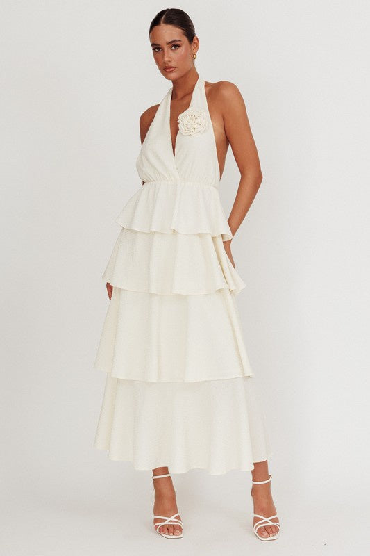 Featuring a halter style tiered maxi dress with a flower detail on the chest in the color beige 