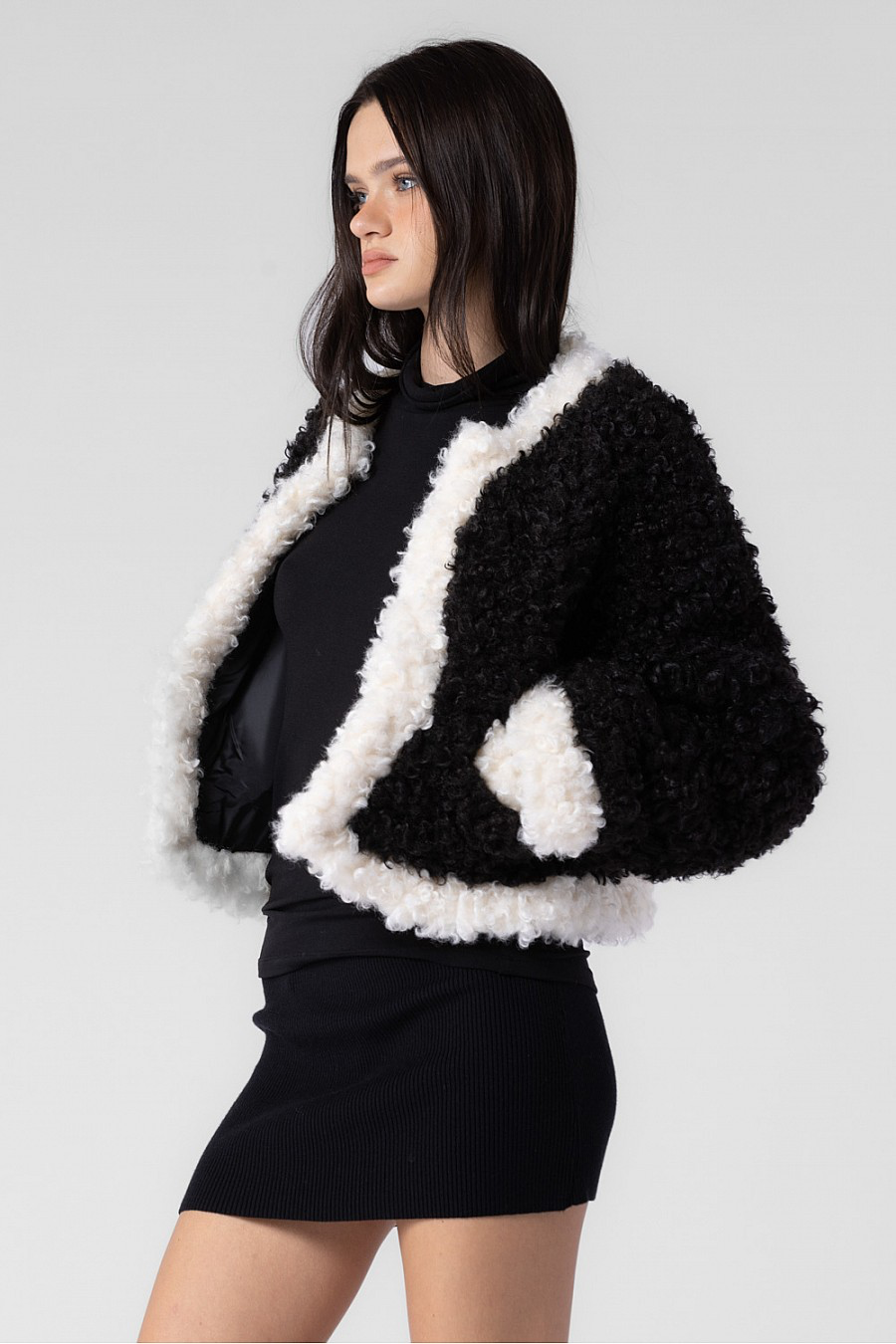 The model is wearing a cropped fluffy jacket with two front pockets. The jacket is black with white lining around the body and cuffs.